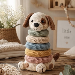 May include: A handmade crochet stacking toy shaped like a dog. The dog has a cream-colored head and body with brown ears and a spot. The toy includes colorful stacking rings in green, beige, blue, yellow, and pink. The toy is on a wooden surface.