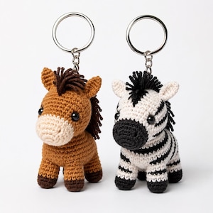 Puede incluir: Dos llaveros de ganchillo con forma de caballo marrón y cebra blanca y negra. Cada animal tiene cabeza redonda, ojos de plástico oscuro y crines cortas. Las figuras están unidas a anillas de metal plateado con pequeñas cadenas.