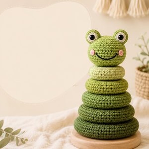 May include: A crochet stacking toy featuring a green frog head topper with black and white eyes and pink cheeks. The toy consists of four circular rings in varying shades of green, stacked on a round, light-colored wooden base stand.