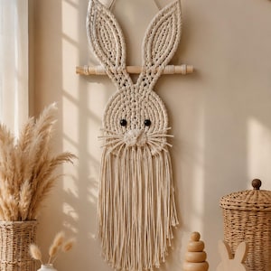 May include: A wall hanging made of off-white macrame cord shaped like a rabbit head with long ears, black bead eyes, and a nose. The design features a wooden dowel at the top for suspension and a long, fringed tassel hanging from the bottom.