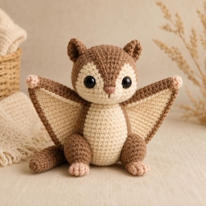 May include: A crocheted flying squirrel toy featuring a brown and cream colour scheme. The plush has large black safety eyes, a small pink nose, and extended wing-like flaps between its arms and body, with light pink accents on its paws.