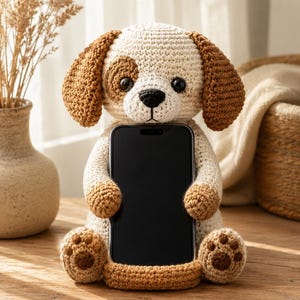 May include: A crocheted dog figurine designed as a phone holder. The dog has cream-colored fur, brown floppy ears, a brown patch over one eye, and a black nose. It is holding a black smartphone upright between its arms and sitting on its feet.