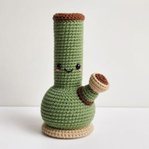 May include: A green crochet water pipe plush with a brown rim at the top and base. The cylindrical body features a smiling face with black bead eyes and pink stitched cheeks. A smaller, angled brown and beige crochet attachment extends from the side.