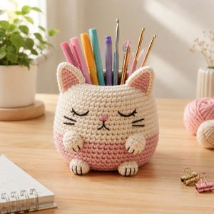 May include: A cream and light pink crocheted cat-shaped desk organizer featuring embroidered closed eyes, whiskers, and a small pink nose. The container holds various colorful pens, pencils, and metal crochet hooks in its open top.