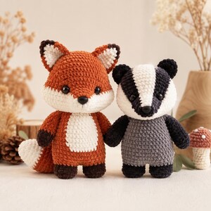Puede incluir: Dos peluches tejidos a mano en crochet que representan un zorro y un tejón. El zorro es naranja con pecho, cara y punta de la cola blancos, y patas marrón oscuro. El tejón es gris y blanco con orejas y patas negras. Ambos tienen ojos y nariz negros.