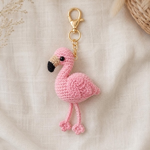 May include: A crochet flamingo keyring featuring a pink yarn body, a beige and black beak, and two small black eyes. The flamingo is attached to a gold-toned metal lobster clasp and a circular ring, providing a secure way to attach the item.