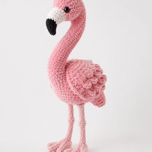 Puede incluir: Una figura de flamenco tejida a ganchillo a mano con hilo rosa suave. El ave presenta un cuello largo y curvado, un pico blanco y negro, y dos pequeños ojos de cuentas negras. Se sostiene sobre dos patas rosas finas y texturizadas.