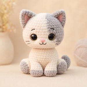Pode incluir: Um peluche de gato feito em croché com uma cabeça de dois tons em fio creme e cinzento. O gato tem grandes olhos de plástico preto, um pequeno nariz bordado a rosa e bigodes bordados. As patas e as orelhas são cinzentas, com detalhes.