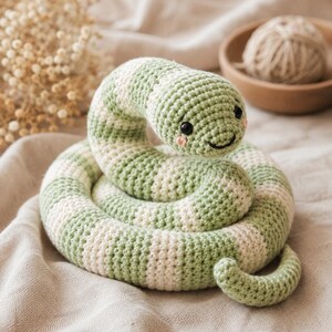 Peut inclure: Un serpent en crochet enroulé avec des rayures horizontales alternées de couleur vert sauge et crème. Le serpent possède deux petits yeux en perles noires, une bouche brodée en noir et deux petites taches roses sur les joues, formant un visage.