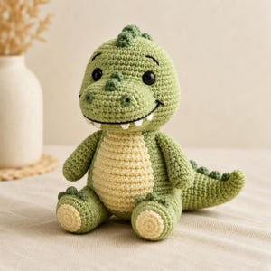 Puede incluir: Un peluche de dinosaurio tejido a ganchillo con cuerpo verde claro, vientre color crema y detalles en verde oscuro a lo largo de la espalda y la cabeza. El juguete tiene ojos de plástico negro, una sonrisa bordada y pequeños dientes blancos.