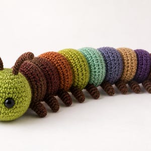 May include: A handmade crochet caterpillar toy in various colors. The caterpillar has a green head with black eyes and a smiling mouth. The body segments are in shades of brown, orange, yellow, green, blue, tan, and purple. The toy is made of yarn.