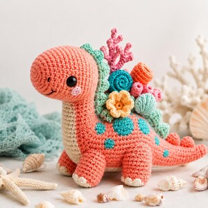 May include: A coral-colored crochet dinosaur with teal spots and a cream-colored belly. The dinosaur has a friendly face and is adorned with crochet coral, flowers, and sea shells in shades of pink, orange, and teal. The background includes seashells and a starfish.