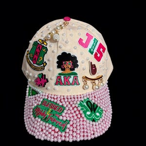 May include: A cream-colored baseball cap adorned with white pearls, featuring embroidered patches including the text AKA, 1908, and Blessed & Highly Favored. The cap includes a central graphic of a person with an afro, a gold-toned pin with dangling pearls, and a green and pink color scheme throughout the various decorative elements.