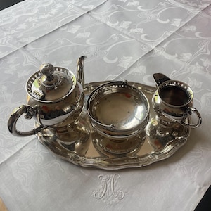 Op de afbeelding: Een zilveren theeservies bestaande uit een theepot, suikerpot en melkkan, gepresenteerd op een zilveren dienblad. De set staat op een witte tafelkleed met een bloemenpatroon en een geborduurd initiaal.