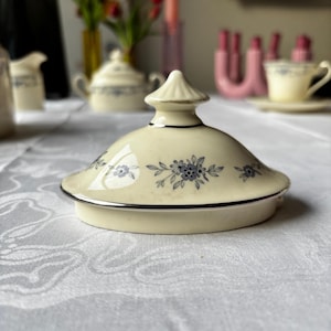 Puede incluir: Tapa de porcelana color crema con borde plateado y un estampado floral azul. La tapa tiene un remate cónico y probablemente forma parte de un juego de té. El fondo está borroso, sugiriendo una mesa.