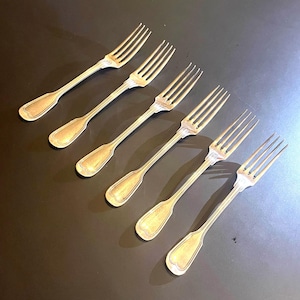 Pode incluir: Um conjunto de cinco garfos de metal prateado dispostos numa fila diagonal. Cada garfo apresenta uma cabeça de quatro dentes e um cabo longo e fino com uma extremidade arredondada e ligeiramente alargada com um rebordo em relevo.