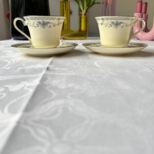 May include: Two cream-colored teacups and saucers with blue floral patterns and gold trim are displayed on a white tablecloth. The teacups have curved handles and sit on matching saucers. The background includes vases and flowers.