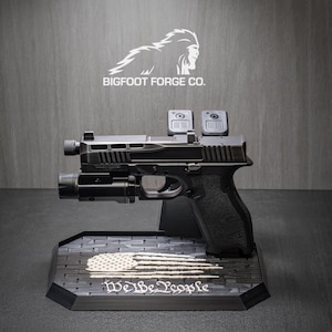 May include: A black handgun with a mounted light and two red dot sights, displayed on a stand. The stand features a stylized American flag design and the text "We the People." The Bigfoot Forge Co. logo is visible above.
