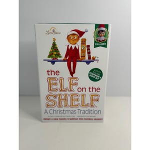 May include: A white rectangular box for The Elf on the Shelf: A Christmas Tradition. The front features an illustration of a scout elf in a red suit sitting on a blue shelf next to a Christmas tree. Text includes the title and I'm a BOY badge.