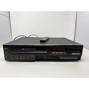 Könnte beinhalten: Ein schwarzer VHS-Videorekorder mit einem Frontlader-Kassettenschacht, einem digitalen Anzeigefeld und zahlreichen Bedientasten. Das Gerät verfügt über eine kleine schwarze Fernbedienung auf der Oberseite sowie verschiedene Anschlüsse.