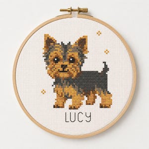 May include: A cross-stitch embroidery in a round wooden hoop featuring a tan and black Yorkshire Terrier dog. The design includes small yellow star accents and the name LUCY stitched in black capital letters on white fabric.