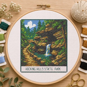 Pode incluir: Uma bordado de ponto cruz concluído do Hocking Hills State Park, apresentando uma cachoeira e vegetação exuberante. A obra de arte é emoldurada em um bastidor de madeira, com carretéis de linha e uma almofada de alfinetes por perto. O texto "HOCKING HILLS STATE PARK" está na parte inferior.