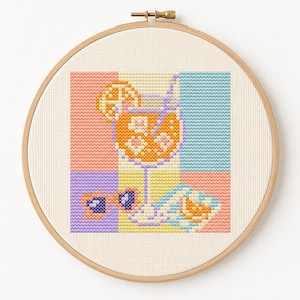 Könnte beinhalten: Eine Kreuzstichstickerei in einem runden Holzrahmen, die ein buntes Cocktailglas mit Orangenscheibe, Eiswürfeln und Strohhalm sowie herzförmige Sonnenbrillen vor einem Hintergrund aus pastellfarbenen rosa, blauen und violetten Quadraten zeigt.