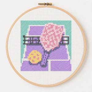 Könnte beinhalten: Eine Kreuzstichstickerei in einem runden Holzrahmen. Das Motiv zeigt einen rosa Pickleball-Schläger, einen gelben Pickleball mit rosa Punkten und ein schwarzes Netz vor einem geometrischen Hintergrund in Violett und Hellblau.