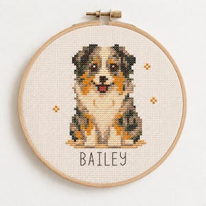 May include: A cross-stitch embroidery in a round wooden hoop featuring a multi-colored Australian Shepherd dog with a white chest and face markings. The name BAILEY is stitched in black capital letters below the dog, with small orange stars.