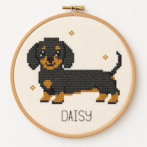 Könnte beinhalten: Ein runder Holzstickrahmen mit einem Kreuzstichmuster eines schwarz-braunen Dackels. Der Hund ist auf weißem Stoff zentriert und von kleinen goldenen Sternen umgeben. Der Name DAISY ist in schwarzen Großbuchstaben unter dem Hund gestickt.