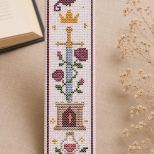 May include: A rectangular cross-stitch bookmark on white fabric featuring a vertical arrangement of a dark purple dragon, a yellow crown, a sword with a blue blade, pink roses, a tan stone tower, a potion bottle, and a yellow key.