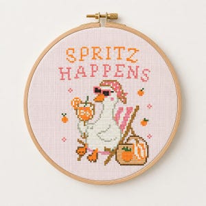 Könnte beinhalten: Eine Kreuzstichstickerei in einem runden Holzrahmen zeigt eine weiße Gans mit rosa Sonnenbrille und gemustertem Kopftuch. Die Gans hält einen orangefarbenen Cocktail. Der Text lautet SPRITZ HAPPENS in orangefarbenen und rosa Buchstaben.