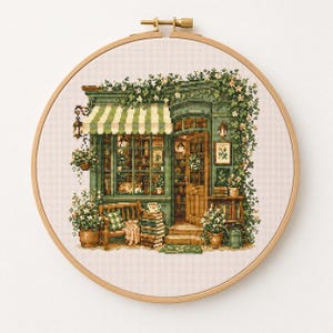 Puede incluir: Bordado de punto de cruz de una pintoresca tienda verde con un toldo a rayas, rodeada de flores y vegetación. La tienda presenta una puerta de madera, escaparates y un banco. El bordado está en un aro de madera.