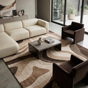 May include: A large area rug featuring a wavy, abstract pattern in shades of cream, beige, and dark brown. The rug is positioned under a rectangular gray coffee table, a cream-colored sectional sofa, and two dark brown upholstered armchairs.