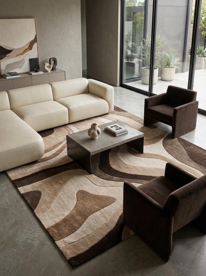 May include: A large area rug featuring a wavy, abstract pattern in shades of cream, beige, and dark brown. The rug is positioned under a rectangular gray coffee table, a cream-colored sectional sofa, and two dark brown upholstered armchairs.