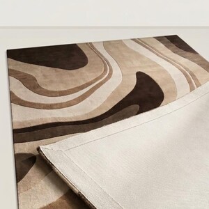 May include: A rectangular area rug featuring a flowing, abstract pattern of wavy lines in shades of dark brown, medium brown, tan, and cream. The rug is partially folded over to reveal a plain, light beige woven fabric backing with stitched edges.