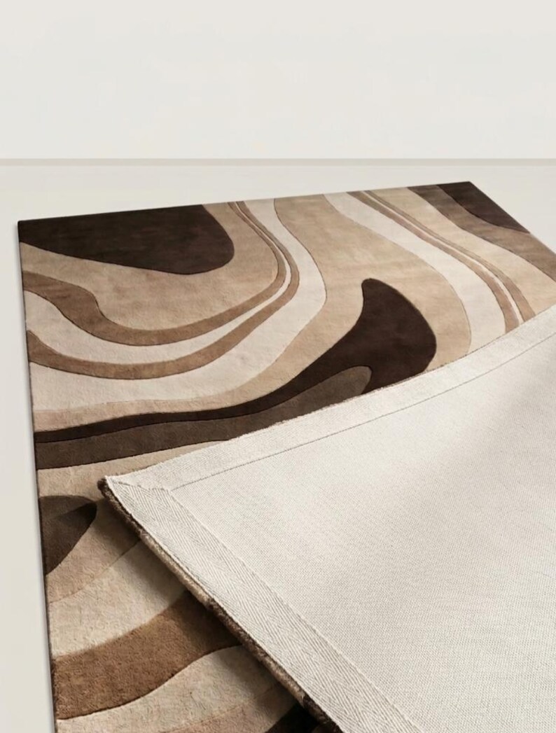 May include: A rectangular area rug featuring a flowing, abstract pattern of wavy lines in shades of dark brown, medium brown, tan, and cream. The rug is partially folded over to reveal a plain, light beige woven fabric backing with stitched edges.