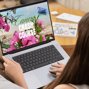 Op de afbeelding: Een zilveren laptop toont een scherm met de tekst "Canva Crash Course" over een strandscène met roze bloemen en een blauwe boot. De laptop staat op de schoot van een persoon.