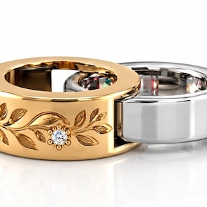 May include: A two-piece interlocking ring set featuring a gold band with an engraved leaf pattern and a small embedded round gemstone, connected to a smooth, polished silver band. The rings are joined together in a linked, circular design.