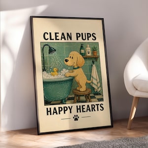 Op de afbeelding: Een ingelijste print met een cartoonhond die een bad neemt in een turquoise badkuip met een badeend. De tekst "CLEAN PUPS HAPPY HEARTS" staat boven en onder de illustratie, met een voetafdruksymbool. De print zit in een zwart frame.