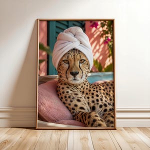 May include: A framed wall art print featuring a cheetah with a white towel wrapped around its head like a turban. The cheetah is resting against a soft pink cushion, with a blurred outdoor setting of green foliage and pink flowers in the background.