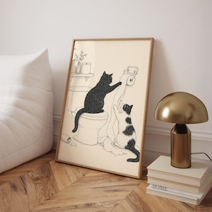 Könnte beinhalten: Gerahmter Kunstdruck mit zwei Katzen in einem Badezimmer. Eine schwarze Katze sitzt auf einer Toilette, während eine andere, schwarz-weiße Katze nach dem Toilettenpapier greift. Das Kunstwerk ist im minimalistischen Stil mit hellem beigen Hintergrund.