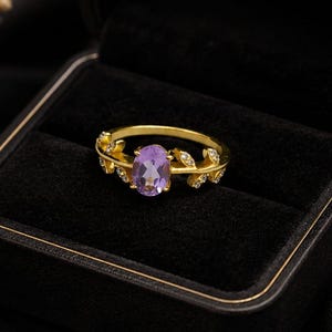 May include: A gold ring featuring an oval-cut purple amethyst gemstone at the center, flanked by delicate gold leaf motifs accented with small clear stones along the band, presented inside a black velvet-lined jewelry box.