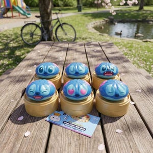 May include: Six blue and pink character-themed fidget toys with different facial expressions, sitting on small, round, yellow containers. A small card with text is visible. The toys are arranged on a wooden picnic table outdoors.