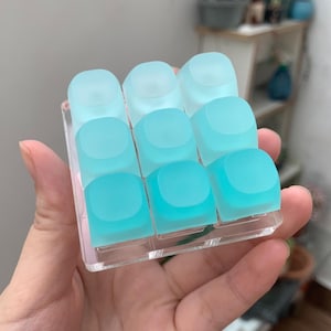 Könnte beinhalten: Ein quadratisches Makro-Pad für mechanische Tastaturen mit neun durchscheinenden, mattierten Tastenkappen in einem Drei-mal-drei-Raster. Die Tastenkappen verlaufen farblich von einem blassen, eisigen Blau oben bis zu einem kräftigen Türkis unten.