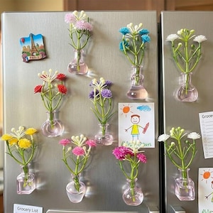 Op de afbeelding: Een verzameling kleurrijke bloemmagneten in kleine glazen vaasjes, bevestigd op een koelkast. De bloemen zijn in verschillende kleuren, waaronder roze, geel, wit en blauw. Een magneet van de Eiffeltoren en een kindertekening zijn ook zichtbaar.