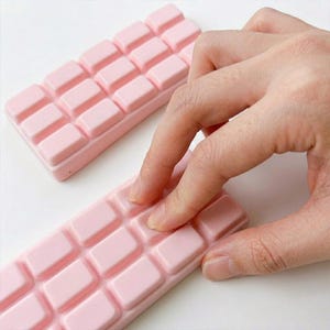 May include: Two pink rectangular bars with a grid pattern, resembling chocolate bars. One bar is in focus, with a person's fingers touching the surface. The bars are on a white surface.