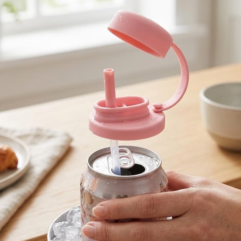 May include: A pink can lid with a straw is placed on top of a silver can. The lid has a hinged cover and a straw that extends into the can. The can is held by a person's hand. A plate with a pastry and a bowl are in the background.
