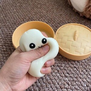 May include: A white, squishy dumpling toy with black eyes and a smiling mouth, held in a hand. Beside it are two halves of a tan, round container, one with a woven pattern.