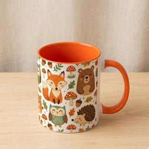 Può includere: Tazza in ceramica bianca con interno e manico arancione. La tazza è decorata con un motivo ripetuto di animali del bosco, tra cui una volpe, un orso, un gufo e un riccio, insieme a funghi e foglie.